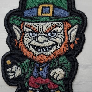 May include: Embroidered patch featuring a leprechaun with a green hat, coat, and shoes, and red pants. The leprechaun has an orange beard and is holding a gold coin. The patch has a black border.
