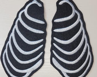 Könnte beinhalten: Zwei schwarze Stoffaufnäher mit weiß gestickten Brustkorbmotiven. Jeder Aufnäher hat eine geschwungene, anatomische Form mit deutlichen weißen Stickereien, die das Aussehen einzelner Rippen auf einem schwarzen Hintergrund nachahmen.