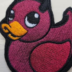May include: Embroidered patch featuring a cartoon duck with devil horns. The duck has a burgundy body, a yellow beak, and black accents. The patch is outlined in black.