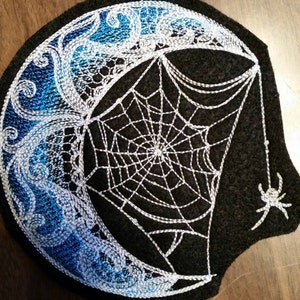 May include: A black felt patch with a white embroidered crescent moon and spider web design. The moon is decorated with blue and white swirls. A small white spider is hanging from the web.