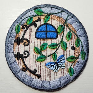 May include: Embroidered patch with a fairy door design. The circular patch features a blue window, green leaves, and a butterfly. The door has a wood-grain texture, and the border has a stone-like pattern.