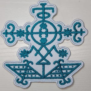 May include: Embroidered patch featuring a teal and white design. The patch has a complex, symmetrical pattern with a cross-like shape, floral accents, and geometric elements. The teal embroidery is set against a white background with a white border.