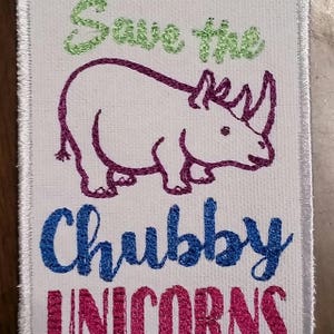 May include: White fabric patch with green, purple, blue, and pink embroidered text that reads "Save the Chubby Unicorns". A purple embroidered rhinoceros is in the center of the patch.