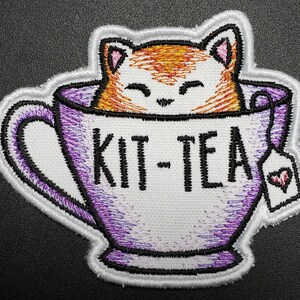 May include: A white teacup with a purple rim and a white tag with a red heart. The teacup has the words "Kit-Tea" written on it. A cartoon orange cat is peeking out of the top of the teacup.