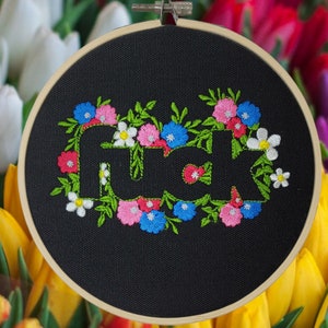 May include: Embroidered hoop art with the word "FUCK" in white, surrounded by a floral wreath. The flowers are in shades of pink, blue, and white, with green leaves, set against a black background.