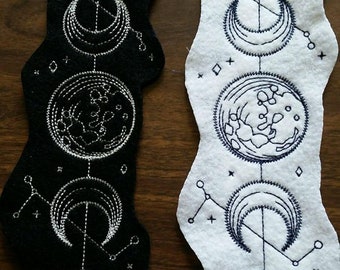 Puede incluir: Dos tiras verticales de fieltro con bordados de fases lunares y constelaciones. Una tira es negra con hilo plateado y la otra es blanca con hilo azul oscuro. Ambas muestran una secuencia de ciclos lunares en un diseño vertical.