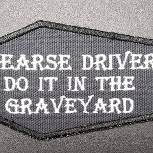 May include: A black patch with white embroidered text that reads "HEARSE DRIVERS DO IT IN THE GRAVEYARD". The patch has a black stitched border and a coffin-like shape.