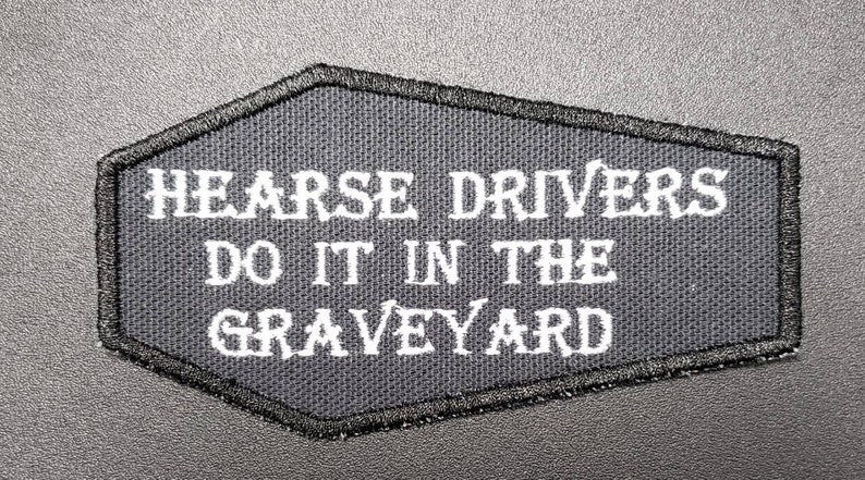 May include: A black patch with white embroidered text that reads "HEARSE DRIVERS DO IT IN THE GRAVEYARD". The patch has a black stitched border and a coffin-like shape.