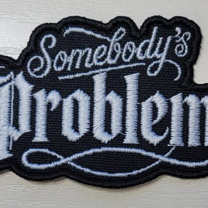 Peut inclure: Un &eacute;cusson brod&eacute; noir avec une bordure cousue blanche et le texte Somebody's Problem dans une police de caract&egrave;res stylis&eacute;e script et serif. La broderie pr&eacute;sente un fond noir avec du fil blanc d&eacute;taillant le lettrage et les fioritures.