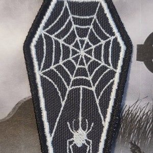 May include: Black and white embroidered patch shaped like a coffin with a spider web and spider inside.