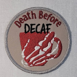 May include: A round, beige fabric patch with a gray border. The patch features a red coffee cup with white skeletal bones and the text "Death Before Decaf" in red and black lettering.