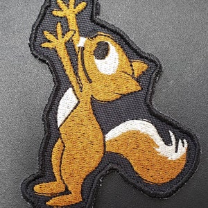 Peut inclure: Un &eacute;cusson brod&eacute; repr&eacute;sentant un &eacute;cureuil de dessin anim&eacute; levant les bras en l'air. L'&eacute;cureuil est debout sur ses pattes arri&egrave;re et a une longue queue touffue. L'&eacute;cusson est brun et blanc.