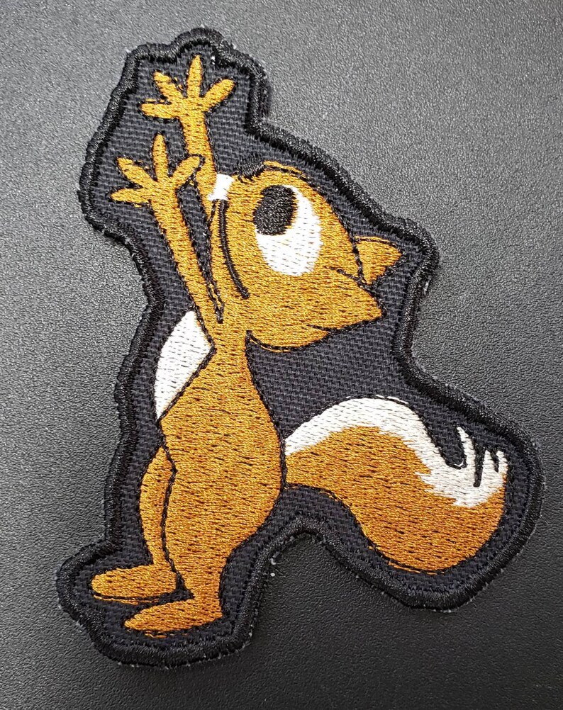 Peut inclure: Un &eacute;cusson brod&eacute; repr&eacute;sentant un &eacute;cureuil de dessin anim&eacute; levant les bras en l'air. L'&eacute;cureuil est debout sur ses pattes arri&egrave;re et a une longue queue touffue. L'&eacute;cusson est brun et blanc.