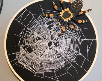 Könnte beinhalten: Ein runder, schwarzer Stoff-Stickrahmen mit einem weißen Spinnennetz-Design. Eine 3D-Spinne mit schwarzen, orangefarbenen und gelben Markierungen ist oben befestigt und schafft eine Halloween-Dekoration.