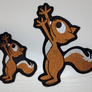 Peut inclure: Deux patchs brod&eacute;s thermocollants repr&eacute;sentant des &eacute;cureuils de dessin anim&eacute;. Le plus grand &eacute;cureuil est brun avec un ventre blanc et se tient debout sur ses pattes arri&egrave;re, les bras lev&eacute;s. Le plus petit &eacute;cureuil est brun avec un ventre blanc et se tient debout sur ses pattes arri&egrave;re, les bras le long du corps.