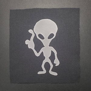 May include: A light gray alien patch on a black fabric square. The alien figure is stylized with large oval eyes and a raised hand. The patch is likely for clothing or accessories.