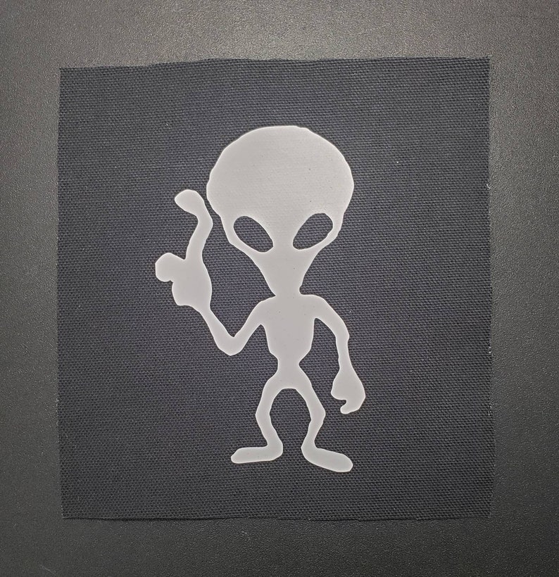 May include: A light gray alien patch on a black fabric square. The alien figure is stylized with large oval eyes and a raised hand. The patch is likely for clothing or accessories.