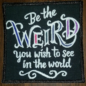 Op de afbeelding: Zwarte geborduurde patch met witte tekst die "Be the weird you wish to see in the world" luidt.