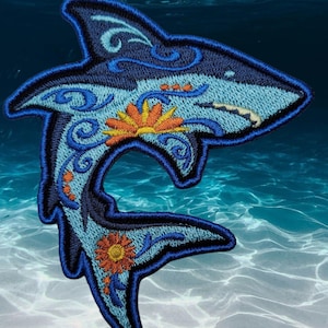 May include: Embroidered shark patch in shades of blue, with orange and yellow floral accents. The patch features intricate detailing and a decorative design, perfect for adding to clothing or accessories. The shark is set against a blue water background.
