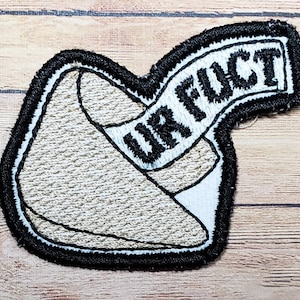 May include: A white and black embroidered patch shaped like a fortune cookie with the text "UR FUCT" on a black banner.