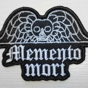 May include: Black embroidered patch with a white skull with wings and the text "Memento Mori" in a gothic font.