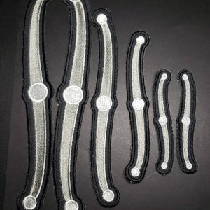 May include: Set of six white and black embroidered patches shaped like bones. The patches are different sizes and are arranged in a row from largest to smallest.