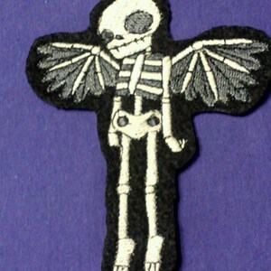 K&ouml;nnte beinhalten: Ein schwarzer Filz-Patch mit einem gestickten wei&szlig;en Skelett mit Fl&uuml;geln. Das Skelett hat einen Sch&auml;del mit einem einzigen Auge und steht mit ausgebreiteten Fl&uuml;geln da.
