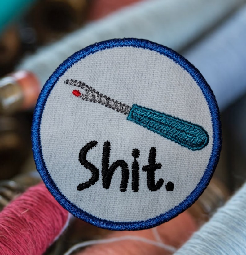 May include: A white embroidered patch with a blue border. The patch features a blue and teal sewing needle with a red eye and the word "shit" in black lettering.