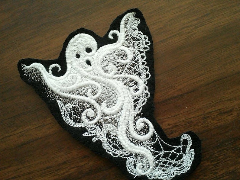 May include: White embroidered ghost with a black background. The ghost is in a lace design with intricate details.
