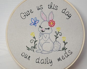 Puede incluir: Arte bordado en bastidor con un conejito, una flor, una mariposa y el texto "Give us this day our daily meds". El diseño incluye flores amarillas y follaje verde, todo bordado sobre tela clara.