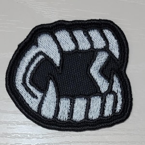 May include: Embroidered patch featuring a stylized set of teeth in black and white. The patch has a black background with white embroidered teeth and a black border. The design is a graphic representation of a mouth with sharp teeth.