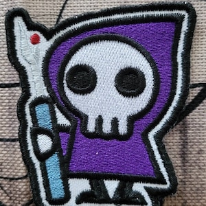 May include: Purple embroidered patch of a cartoon skeleton wearing a purple hooded robe and holding a blue staff. The skeleton has a white skull with black eyes.