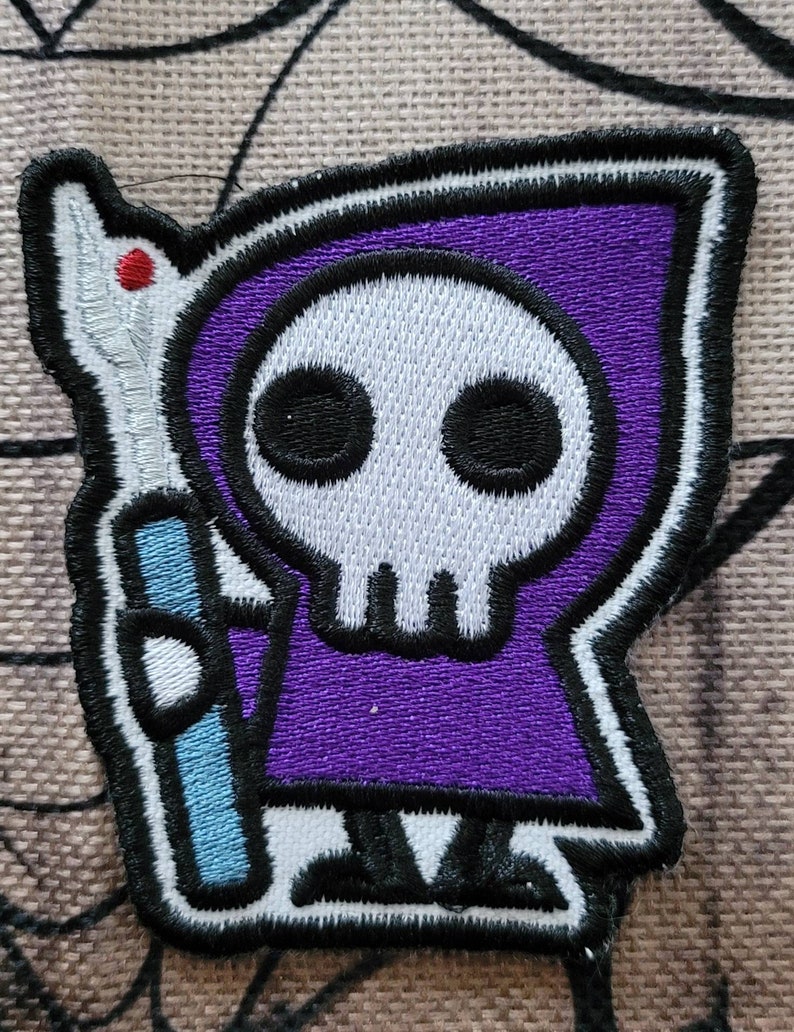 May include: Purple embroidered patch of a cartoon skeleton wearing a purple hooded robe and holding a blue staff. The skeleton has a white skull with black eyes.