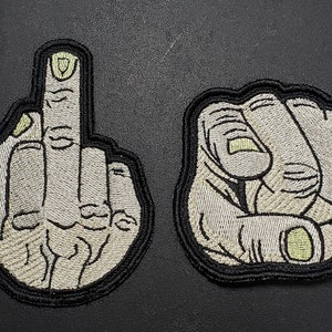 May include: Two embroidered patches. One shows a hand giving the middle finger, the other a clenched fist. Both are in shades of gray with black borders.