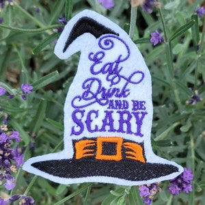 May include: A white witch hat patch with the words "Eat Drink AND BE SCARY" embroidered in purple. The hat has a black brim and a bright orange buckle. The patch is set against a background of green leaves and purple flowers.