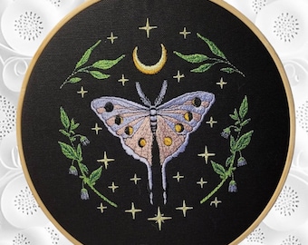 May include: Embroidered hoop art featuring a purple and pink butterfly with yellow accents, surrounded by green leaves and a crescent moon on a black background.