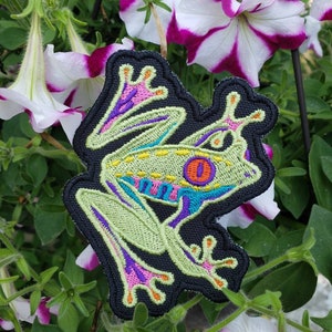 May include: Embroidered patch featuring a vibrant tree frog design. The frog has a light green body, with blue, purple, and orange accents. The patch is outlined in black and set against a backdrop of white and purple petunia flowers.