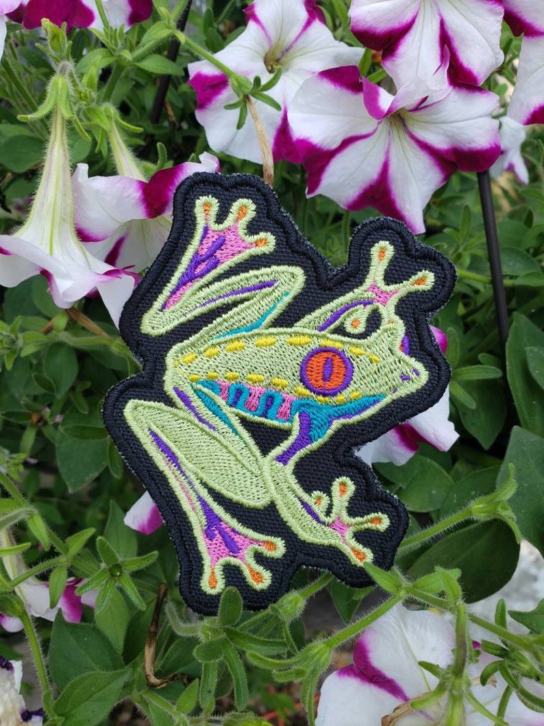 May include: Embroidered patch featuring a vibrant tree frog design. The frog has a light green body, with blue, purple, and orange accents. The patch is outlined in black and set against a backdrop of white and purple petunia flowers.