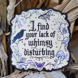 May include: A square embroidered patch with a scalloped black border on a beige background. It features the black text "I find your lack of whimsy disturbing" surrounded by purple and blue embroidered motifs including a raven, butterflies, mushrooms, and vines.