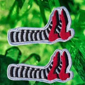 May include: Two embroidered patches featuring the Wicked Witch of the West's striped stockings and ruby red slippers. The patches have a white border and are set against a green background.