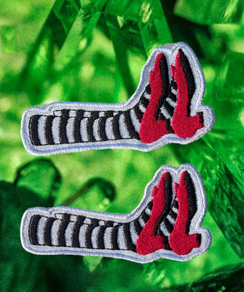 May include: Two embroidered patches featuring the Wicked Witch of the West's striped stockings and ruby red slippers. The patches have a white border and are set against a green background.