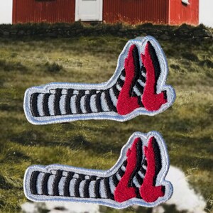 May include: Two embroidered patches featuring a pair of red high-heeled shoes and black and white striped legs. The patches have a light blue border and are set against a blurred outdoor background.