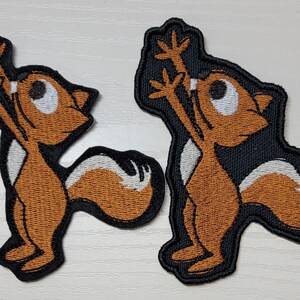 Peut inclure: Deux patchs brod&eacute;s thermocollants repr&eacute;sentant un &eacute;cureuil de dessin anim&eacute; avec une fourrure brune, un ventre blanc et des contours noirs. L'&eacute;cureuil est debout sur ses pattes arri&egrave;re, les bras lev&eacute;s en l'air.