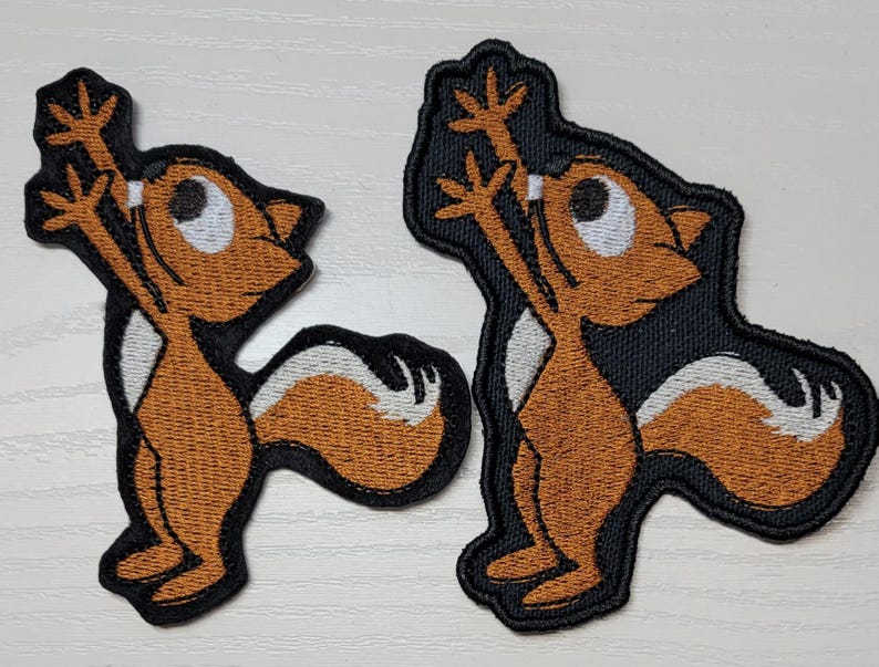 Peut inclure: Deux patchs brod&eacute;s thermocollants repr&eacute;sentant un &eacute;cureuil de dessin anim&eacute; avec une fourrure brune, un ventre blanc et des contours noirs. L'&eacute;cureuil est debout sur ses pattes arri&egrave;re, les bras lev&eacute;s en l'air.