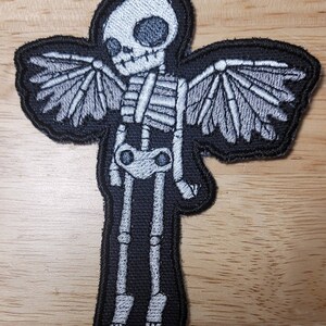 K&ouml;nnte beinhalten: Ein gestickter Patch in Schwarz und Wei&szlig;, der ein Skelett mit Fl&uuml;geln zeigt. Das Skelett tr&auml;gt ein schwarzes Hemd und hat einen wei&szlig;en Sch&auml;del. Die Fl&uuml;gel sind wei&szlig; und haben einen schwarzen Umriss.