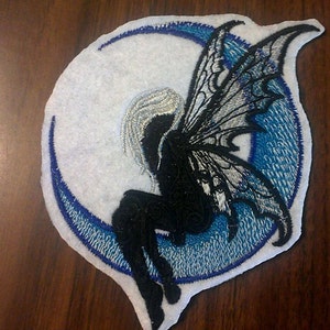 May include: A black and white embroidered patch of a fairy with wings sitting on a blue crescent moon. The patch is on a white background.