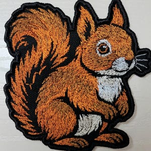 Op de afbeelding: Geborduurde patch met een eekhoornontwerp. De eekhoorn heeft oranje en witte vacht met een zwarte omtrek. Geschikt voor kleding of accessoires.