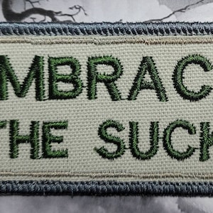 May include: A white embroidered patch with a blue border. The patch says "Embrace the Suck" in green thread.