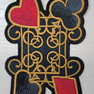 May include: A black fabric patch with a gold embroidered design of a playing card suit. The design features a heart, a spade, and a diamond, all with gold outlines. The patch is perfect for adding a touch of whimsy to clothing, bags, or other accessories.