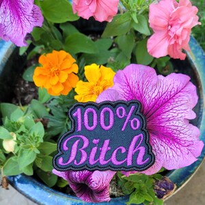 May include: A patch with the text "100% Bitch" in pink lettering on a navy blue background, surrounded by a black border. The patch is placed among vibrant pink and purple petunias, orange and yellow marigolds, and green foliage in a blue ceramic pot.